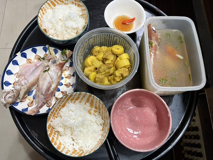 A top-down view of a Japanese meal served on a black tray, featuring two bowls of white rice, a plate of cooked fish, a bowl of yellow curry, a clear soup with fish in a plastic container, a pink empty bowl, and a small dish with a chili.