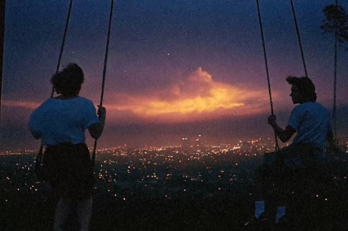 Two people sit on swings at dusk, silhouetted against a glowing purple and orange sky over a city dotted with twinkling lights, evoking a serene, hopeful morning moment that complements the encouraging "Good morning CT, let's win" message.