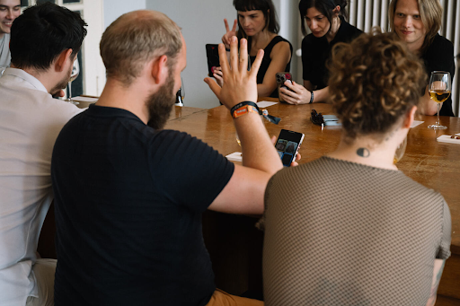 A group of people seated around a wooden table in a meeting, with a bearded man in a black t-shirt raising his hand while another person uses a smartphone, and drinks including beer visible on the table.