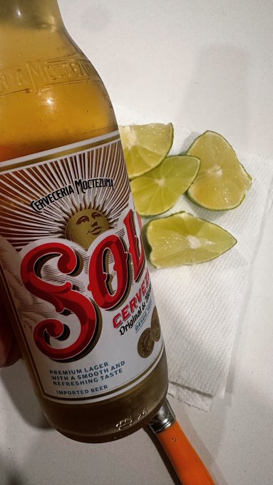 A hand holding a bottle of Sol beer (Cerveceria Moctezuma) next to lime wedges resting on a white paper towel.