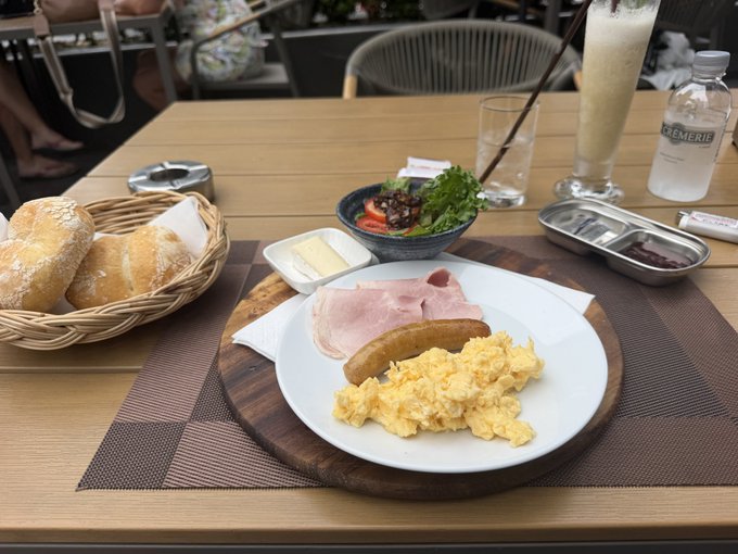 A breakfast spread on a wooden table featuring scrambled eggs, ham, and sausage on a white plate, accompanied by a basket of bread, a small salad, a latte, and a bottle of water.