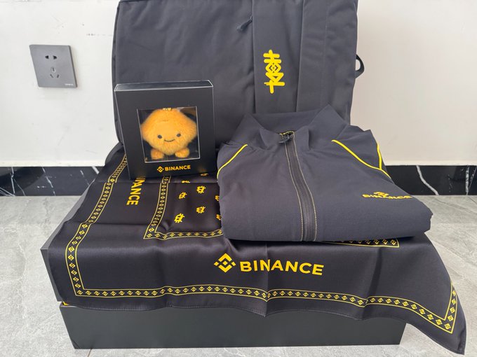 A black gift box from Binance's 9th anniversary contains a plush toy, a jacket, and a bandana, all branded with the Binance logo and the Chinese character for "joy." This imagery directly relates to the post by showcasing the merchandise received for Binance's 9th anniversary.