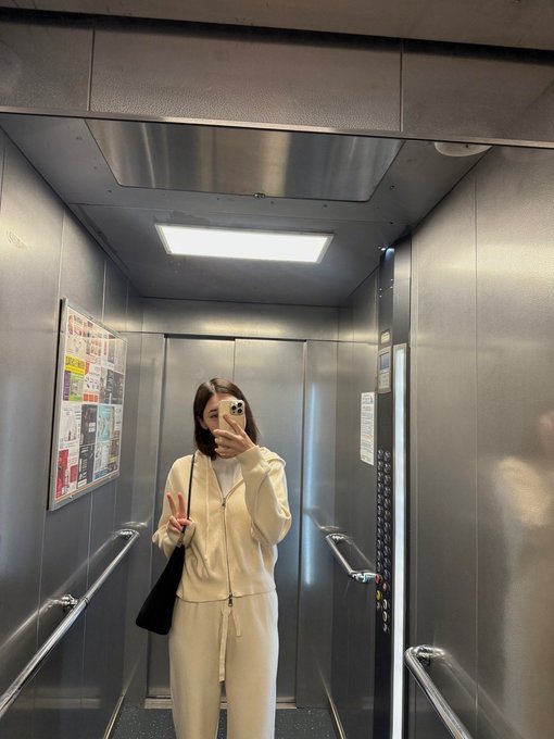 A woman in a cream-colored tracksuit takes a mirror selfie with a peace sign inside an elevator with metallic walls and handrails.