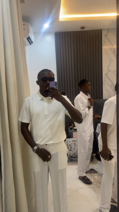 A mirror selfie shows a man wearing sunglasses and a white polo shirt taking a photo with his phone, while other people dressed in white stand in the background.