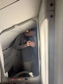 A man taking a mirror selfie inside what appears to be an airplane lavatory, holding a camera while wearing a cap and sunglasses.
