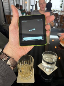 A hand holds a small black device displaying 'Tap mode' on its screen, with a glass of water and amber drink visible on the table below.