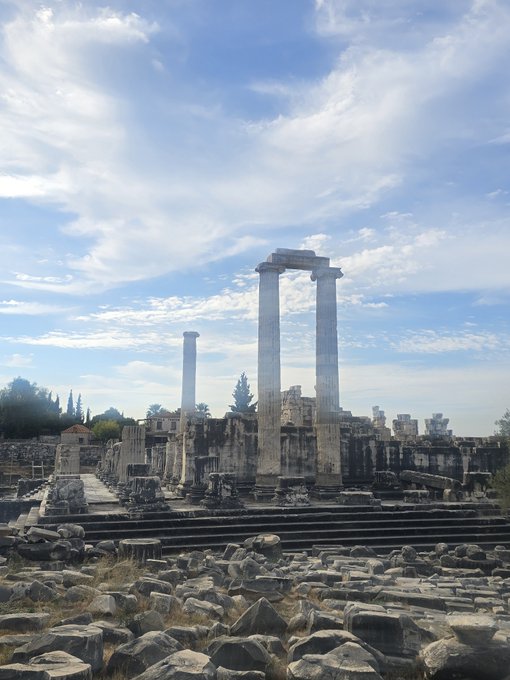Ancient stone ruins stand under a partly cloudy blue sky, featuring a set of standing columns and scattered stone blocks across the site.