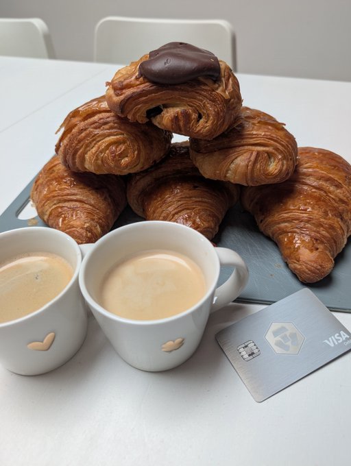 A pile of golden croissants topped with one chocolate-filled pastry sits on a cutting board behind two white espresso cups, accompanied by a silver Visa credit card.