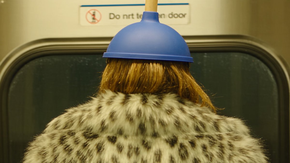 A person wearing a leopard-print fur coat stands with their back to the camera inside a train, with a blue plunger humorously suctioned to their head in front of a door marked 'Do not [lean on] door'.