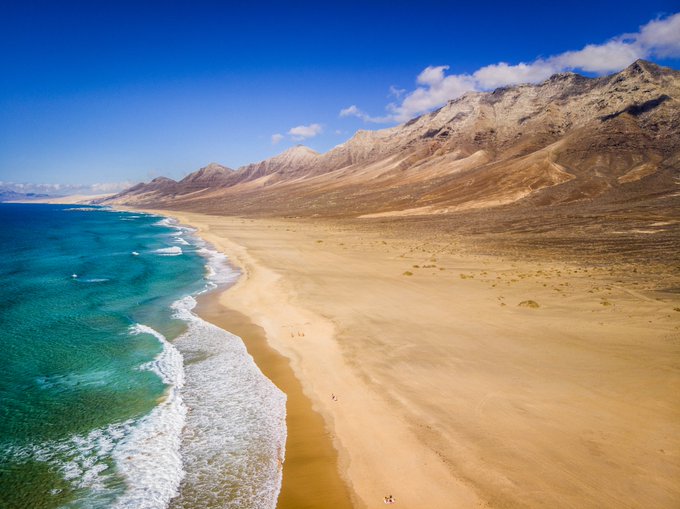 An aerial view of a long sandy beach meeting turquoise ocean waves, with arid mountains rising in the background under a blue sky.