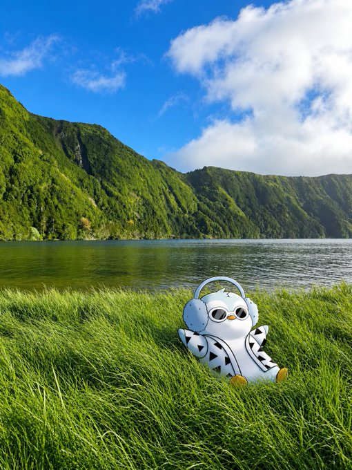 Scenic landscape showing a small white astronaut-like plush figure with headphones sitting in tall green grass beside a lake, with lush green mountains and blue cloudy sky in the background.