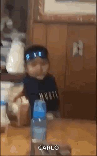 The image features a baby wearing a blue hat, sitting at a dining table with various objects around him. There are two bottles of water placed near his feet, one closer to the left side of the table and another towards the right side. A cup is also present on the table, located in front of the baby's head. The background includes a wooden wall and a chair, suggesting that this scene takes place in a dining area or kitchen setting.