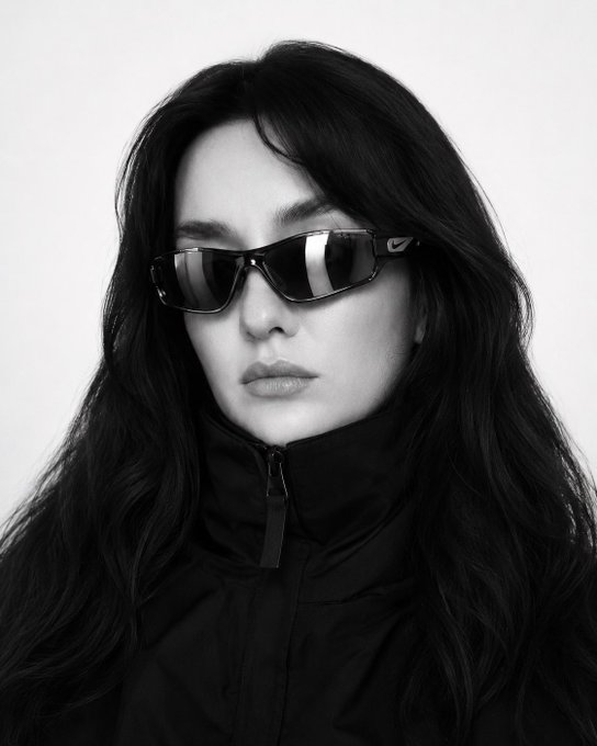 A black-and-white portrait of a woman with long dark hair wearing Nike sunglasses and a black zip-up jacket.