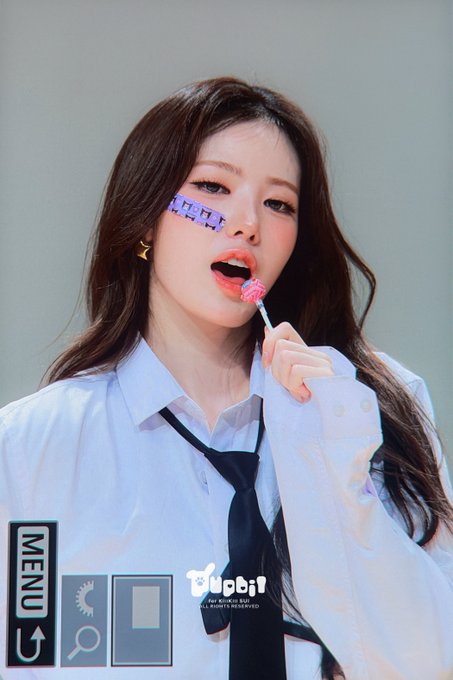 A young woman with long dark hair wears a white collared shirt and black tie, holding a pink lollipop to her lips while sporting purple tape on her cheek that reads "16371." The image features "MENU" text, search icons, and a "KiiiKiii - I FOUND YOU" watermark, matching the post's reference to Sui of KiiiKiii in a pre-debut concept photo.