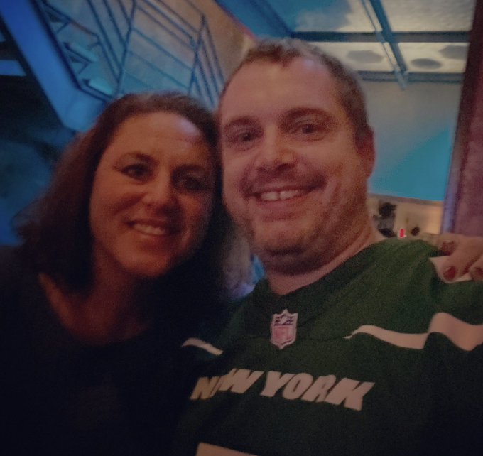 A man wearing a green New York Jets NFL jersey poses for a selfie with a smiling woman in a dimly lit venue.