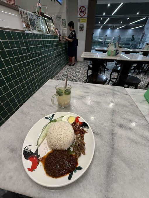 A plate of food featuring white rice and dark red sauce/gravy with garnishes, accompanied by a mug of citrus-infused water, set on a marble table in a casual eatery with green tiled walls.