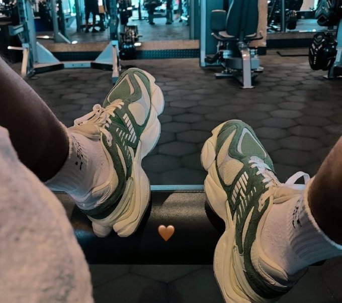 A first-person view of someone wearing green and white chunky sneakers, resting their feet on a piece of gym equipment with a small gold heart emoji between the shoes.