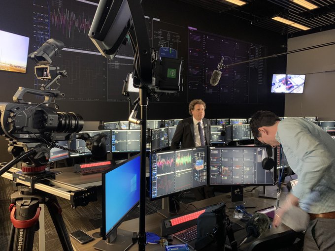 A behind-the-scenes view of a broadcast control room with multiple monitors showing stock charts, a camera operator, and a presenter in a suit preparing for a news segment.