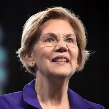 A close-up photograph of a smiling woman with short blonde hair and glasses, wearing a purple blazer against a dark background.