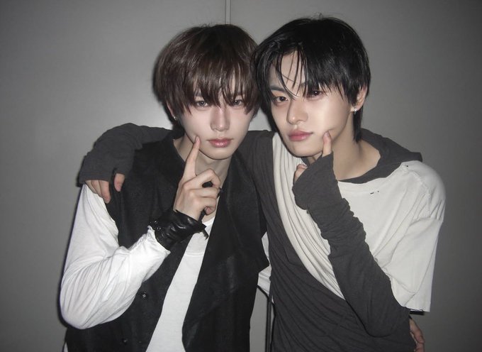 Two young men in monochromatic outfits pose together against a gray background, both pointing fingers near their faces.