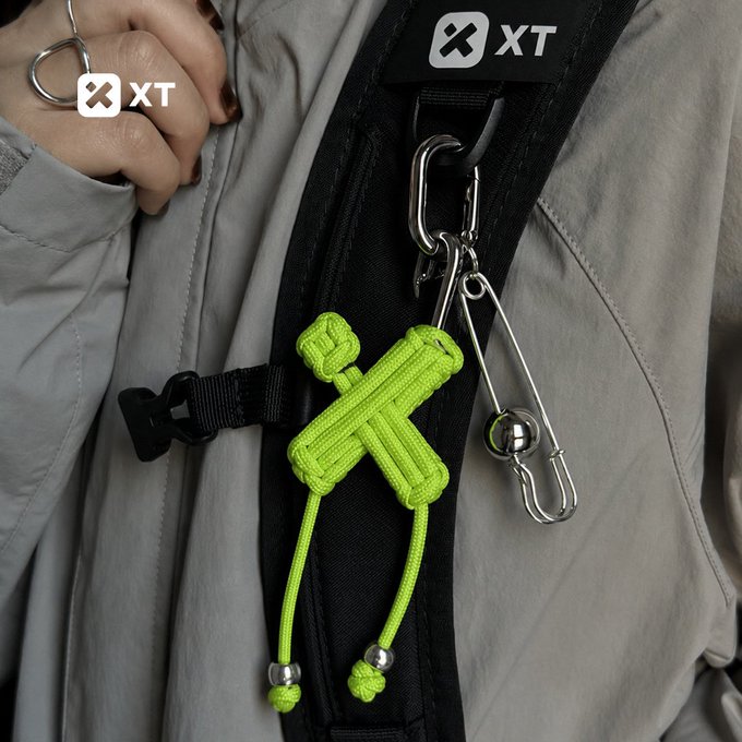 This image shows a neon green "XT" keychain and a safety pin with a ball charm attached to a backpack, accessorized with an "XT" ring on a finger. The post's message about "XT" and "everyday moves" directly relates to the stylish and practical keychain shown.