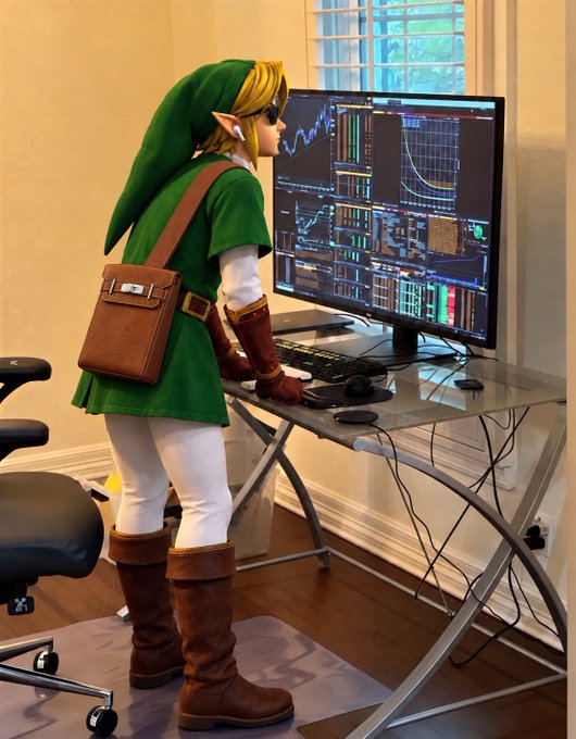 A person dressed as the video game character Link stands at a desk using a computer monitor displaying cryptocurrency trading charts, candlestick graphs, and market panels. The setup matches the post’s joke about being “locked in monitoring the situation” and deciding which coin to watch.