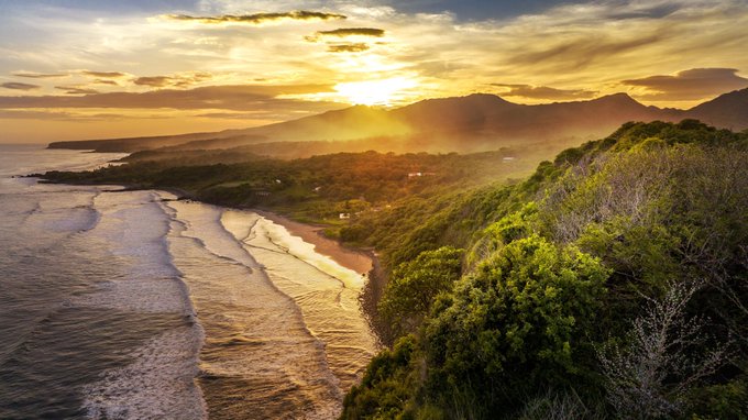 A stunning aerial view captures a sunset over a coastal landscape, with golden light streaming through clouds over a winding beach, lush green hills, and distant mountains.