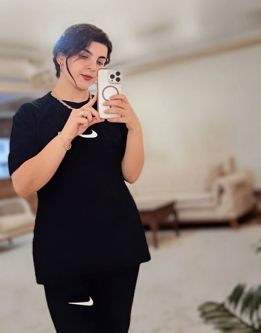 A woman wearing a black Nike t-shirt and matching black Nike pants takes a mirror selfie with her iPhone in a well-lit living room.