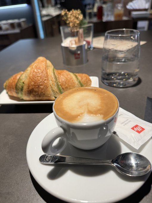 A cappuccino with "illy" branded sugar packet, a croissant, and a glass of water sit on a dark café table, paired with the post "gm legends" as a simple morning ritual.