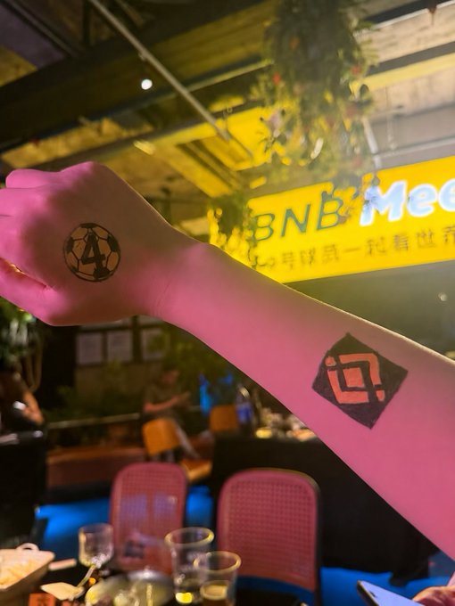 A person's outstretched arm displays two temporary tattoos—one of a soccer ball and another diamond-shaped logo—at what appears to be a 'BNB Meet' event, with a yellow illuminated sign visible in the background.