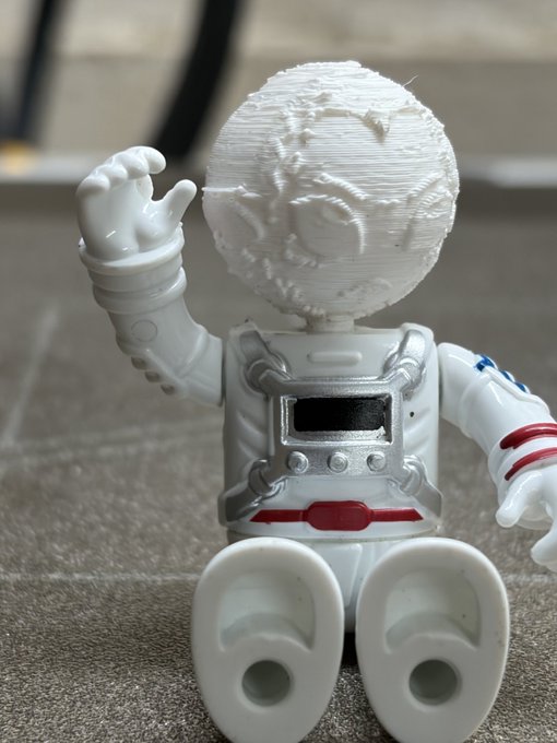 A 3D-printed astronaut figurine with a textured moon-like helmet sits on a surface, with its hand raised in a waving gesture.