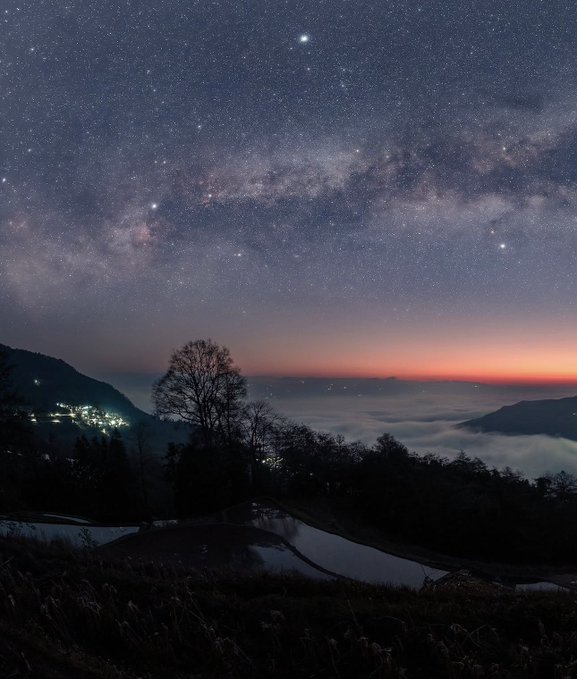 A stunning night sky photograph showing the Milky Way galaxy stretching over a landscape of mountains, clouds, and distant lights, with reflective water pools in the foreground.