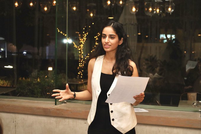 A young woman with long dark hair, wearing a black top and white vest, stands holding papers and speaking at what appears to be a presentation or event, with decorative string lights visible through the window behind her. The image illustrates her journey from running a mountain cafe to speaking publicly, marking her 5-year milestone in the crypto space.