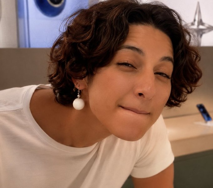A close-up selfie of a smiling woman with short curly brown hair wearing a white shirt and pearl drop earrings.