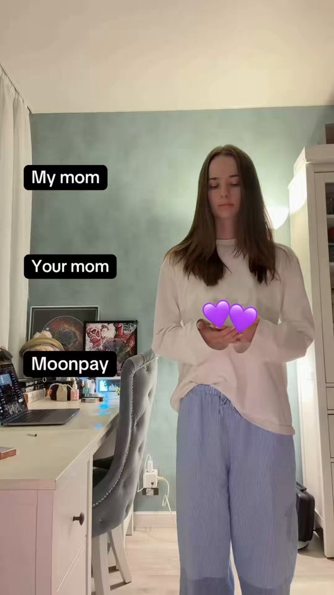 The image features a young woman standing in front of a desk with a laptop, holding up two hearts as if she's about to send them off into space or someone else's heart. The background shows a wall painted green, which contrasts with her white shirt and blue pants. There is also a chair visible behind the woman. The post includes text that reads "My mom Your mom Moonpay it's time for new drop".