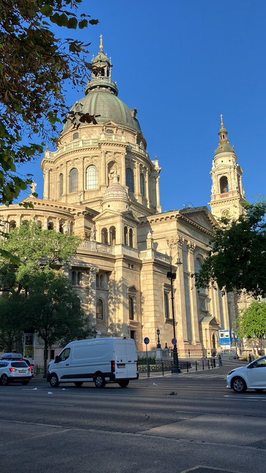 Ornate baroque cathedral with a large dome and bell towers under a clear blue sky, with a white van and cars parked on the street below.