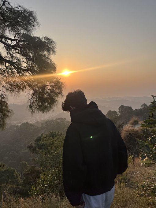 A silhouette of a person in a dark hoodie standing at a scenic overlook, watching the sunrise over misty mountains and pine trees.