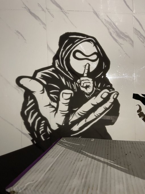 Black and white street art mural of a hooded figure with goggles making a "shh" gesture, painted on a tiled wall.