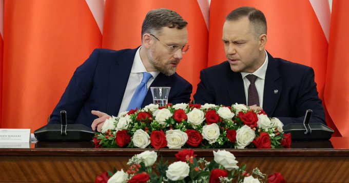 Two men in dark suits converse closely while seated behind a table adorned with red and white roses, with Polish flags displayed prominently in the background. A nameplate in the lower left appears to read "ANDRZEJ BANASZAK," and the scene reflects the post's political context, suggesting this image captures Polish officials—including likely figures from President Duda's office—engaged in a discussion, contextualized by the caption's criticism of their veto of cryptocurrency regulation and subsequent blame directed at Donald Tusk's government.
