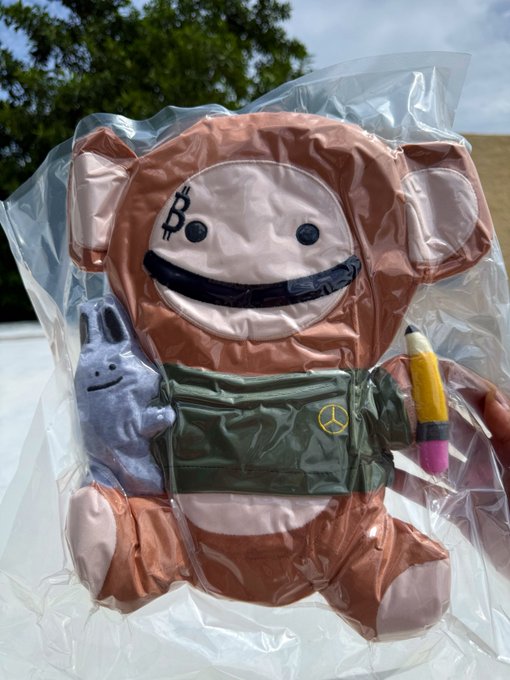 A plush toy in plastic packaging featuring a brown monkey-like character with a Bitcoin 'B' logo on its forehead, holding a green backpack, a small blue bunny, and a pencil.