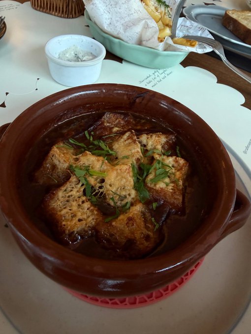 A brown clay pot filled with a dark stew topped with toasted bread cubes and fresh parsley, served alongside a small bowl of white sauce.