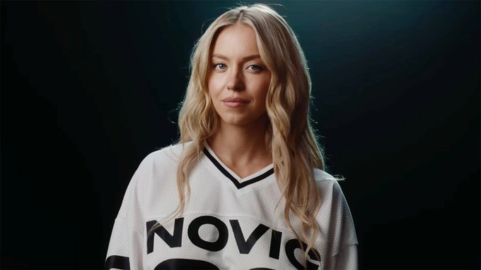 A blonde woman poses in a white jersey with 'NOVIC' text visible, photographed against a dark background with professional lighting.