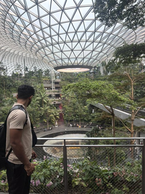 A man with a backpack stands looking over a railing at the interior of Jewel Changi Airport in Singapore, featuring a large indoor waterfall and lush vegetation under a geometric glass dome.