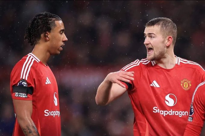 Two Manchester United football players in red Adidas jerseys with the Snapdragon sponsor logo appear to be in a heated discussion on the pitch, with the captain's armband visible on one player's sleeve.