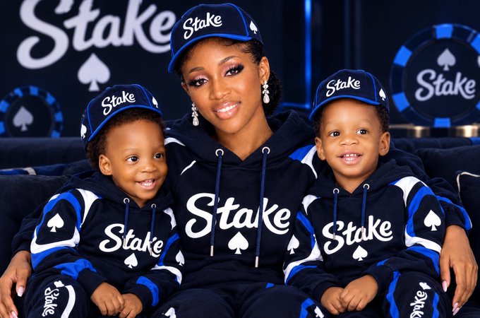 A photograph of a smiling woman seated with two young boys, all wearing matching dark blue 'Stake' branded hoodies and caps, set against a poker-themed backdrop.