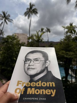 A hand holding the book 'Freedom Of Money' by Changpeng Zhao, with a tropical background featuring palm trees and a pool.