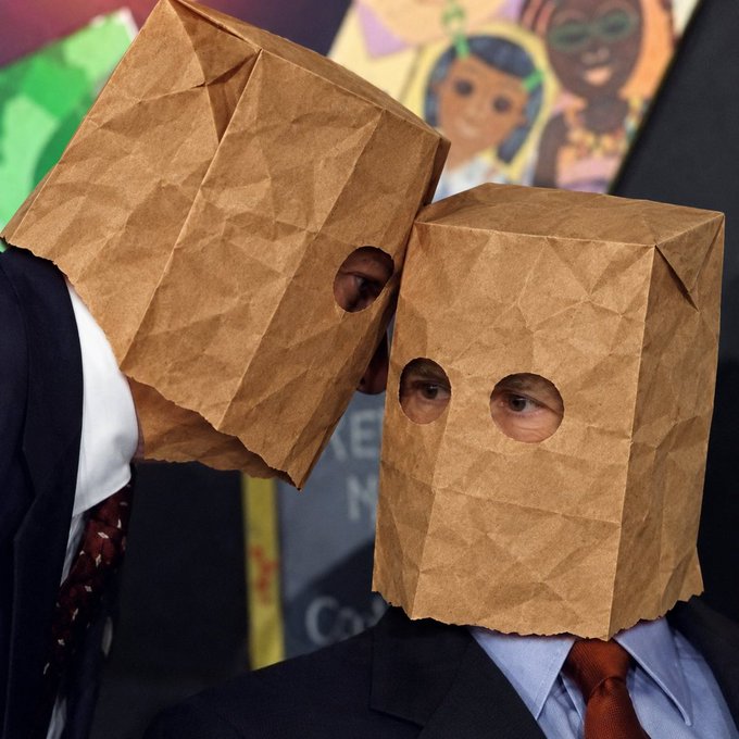 Two people wearing business suits and ties have brown paper bags with eye holes over their heads, standing in front of a colorful cartoon poster.