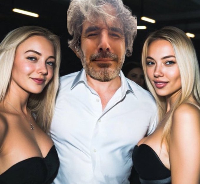 A man with gray hair and a beard in a white shirt poses between two blonde women wearing black outfits, all smiling at the camera.