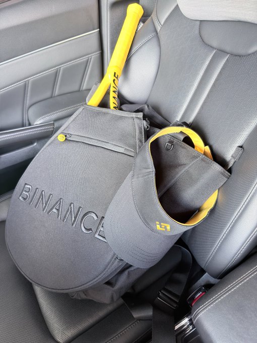 A gray backpack with 'BINANCE' embossed on it and a yellow baseball bat with 'BINANCE' printed on it rest on the black leather seat of a car.
