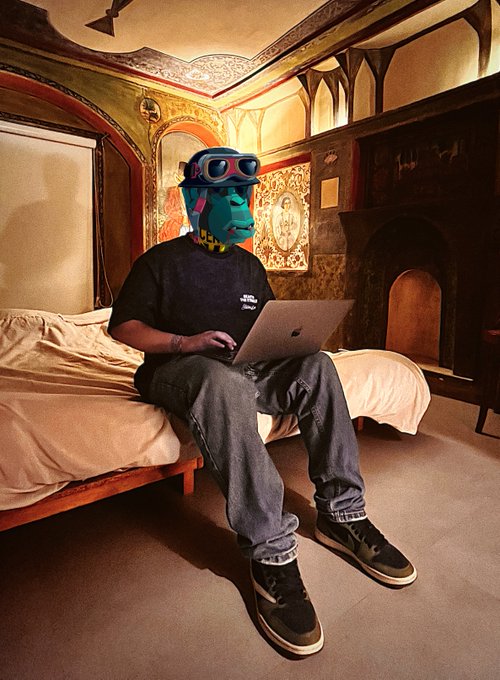 A person wearing a teal mask with VR goggles, sitting on a bed in an ornate, cave-like room while using a laptop.